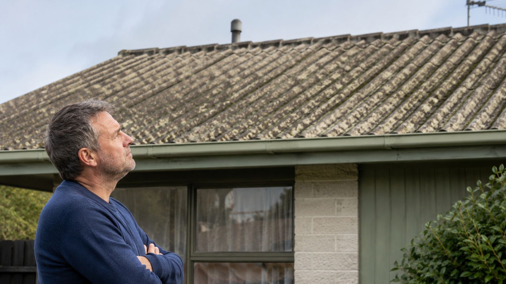 En midaldrende mand kigger bekymret op på et gammelt og beskidt eternittag med asbest på et hus.